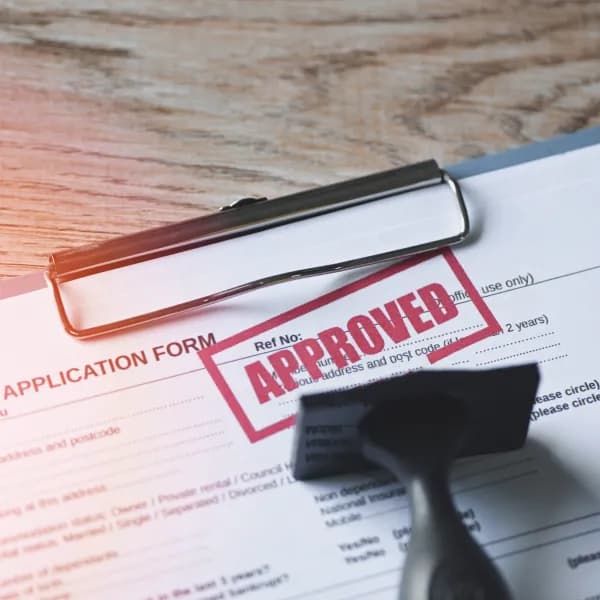 A paper application form on a clipboard, marked with a red "APPROVED" stamp, with the rubber stamp in the foreground.